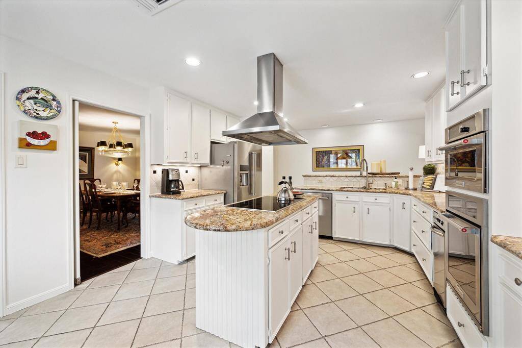 This spacious kitchen features modern stainless steel appliances, granite countertops, and ample white cabinetry. The central island with a cooktop and vent hood offers functionality, while the open layout connects seamlessly to a formal dining area. Ideal for entertaining and family cooking.