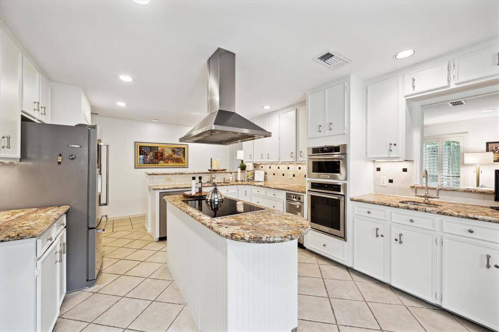 This kitchen features a spacious, modern design with white cabinetry, granite countertops, and stainless steel appliances. An island with a cooktop and stylish range hood enhances functionality, while ample lighting and tiled flooring add to the bright, inviting atmosphere.