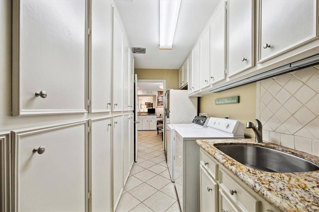 This photo shows a well-organized laundry room with ample cabinetry for storage, a countertop with a sink, and a washer and dryer. The space features neutral tones and tiled flooring, offering functionality and convenience.