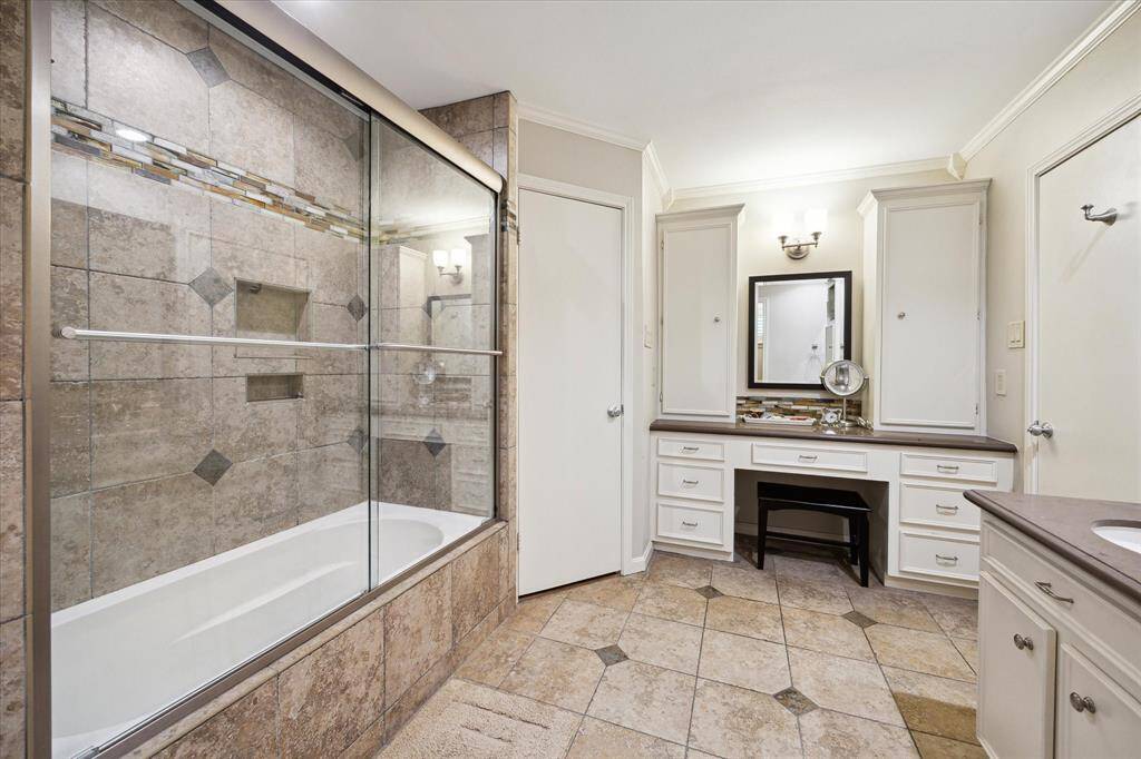 This bathroom features a spacious layout with a tiled bathtub and shower combo, large vanity with ample storage, and elegant lighting. Neutral tones and built-in cabinetry provide a modern and functional space.