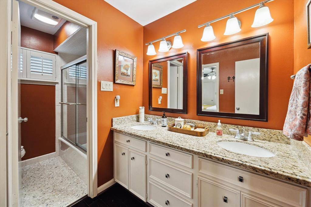 This bathroom features a dual-sink vanity with granite countertops and ample cabinetry. It has warm, orange walls complemented by bright lighting fixtures above the mirrors. The shower area is enclosed with sliding glass doors, and there's a window with shutters for natural light.