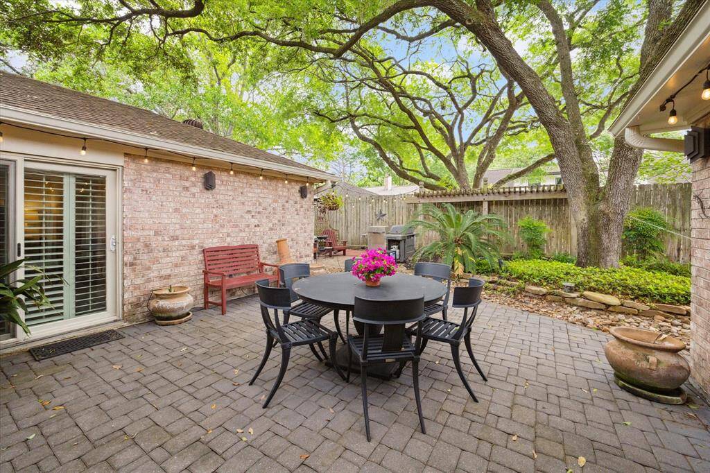 Charming backyard patio with a pavers, featuring a cozy dining area and lush greenery. Mature trees provide shade, while string lights add ambiance. Ideal for outdoor entertaining and relaxation.