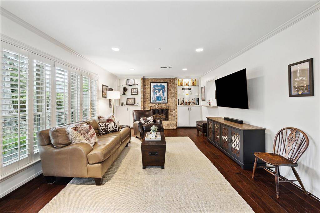 Bright and inviting living room with large windows and shutters, wood flooring, and a cozy brick fireplace. The space features a comfortable seating area, built-in shelving, and a wall-mounted TV. Perfect for relaxation and entertainment.