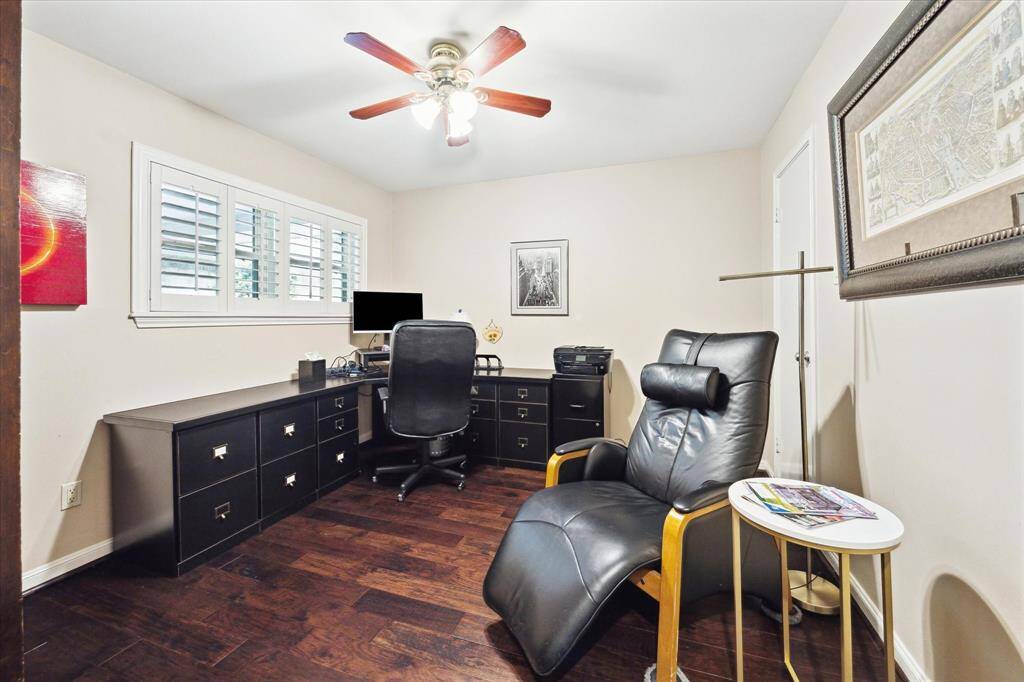 Bedroom being used as a home office with wood flooring. The room is well-lit by a ceiling fan with lights and a window with shutters, offering a bright and inviting workspace with views of the tree lined street