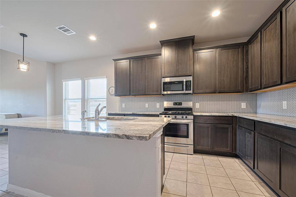 Thoughtfully designed for both function and flow, the kitchen features rich upper and lower cabinetry, expansive granite countertops, and a textured backsplash that adds visual interest. The central island offers seating, a sink, and extra storage—all under soft recessed lighting.