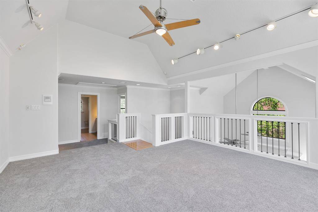 Bright and spacious loft area with vaulted ceilings, track lighting, and a ceiling fan. Features a carpeted floor and stylish railing overlooking a large arched window.
