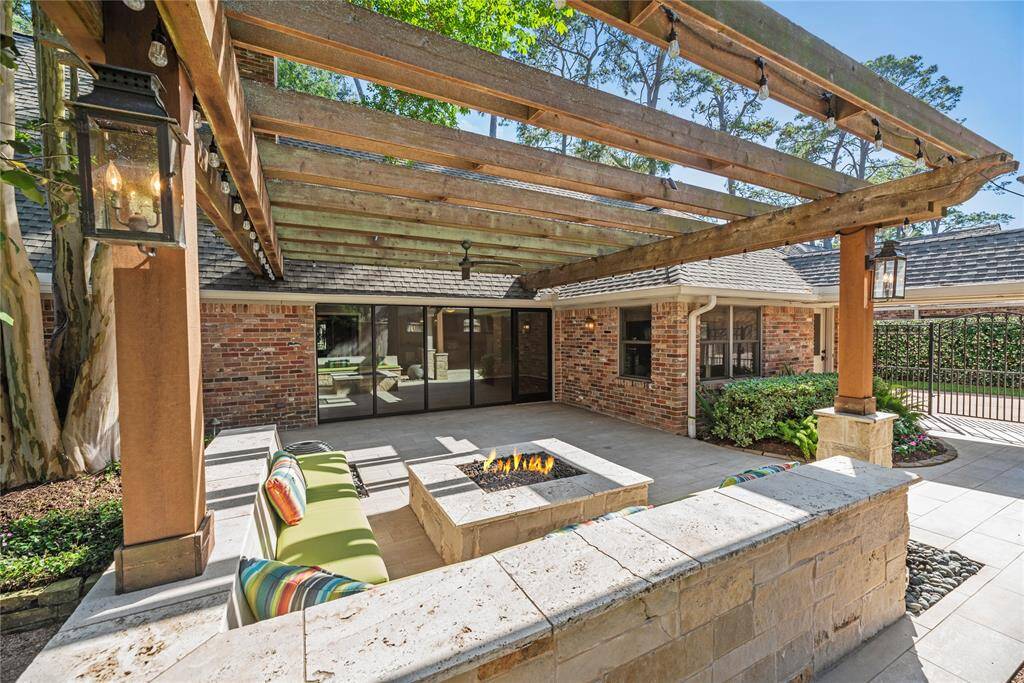The backyard lounge area with a stone fire pit, wraparound cushioned seating is framed by a rustic wood pergola with festoon lighting—ideal for gatherings or quiet evenings.