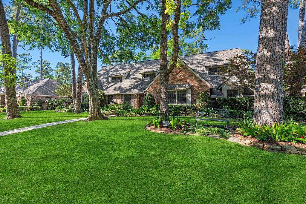 A wide-angle side view of the lush front yard and the home’s beautiful façade. The landscaping features vibrant greenery and a cozy sitting bench nestled under towering trees.