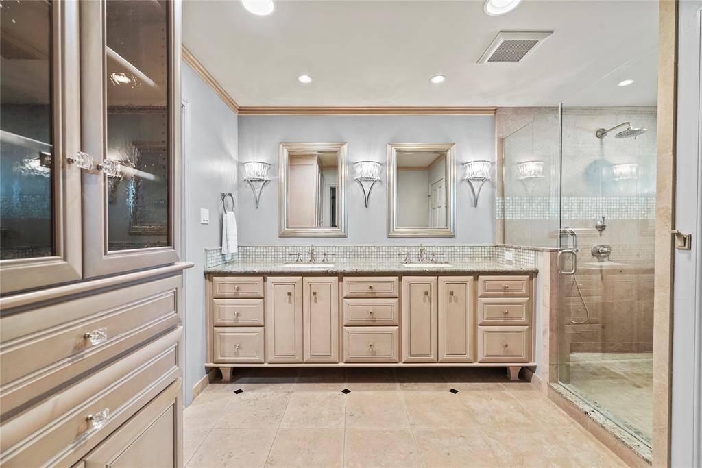 This luxurious ensuite bath includes dual vanities with granite countertops, framed mirrors, designer sconce lighting, and a seamless oversized walk-in shower.