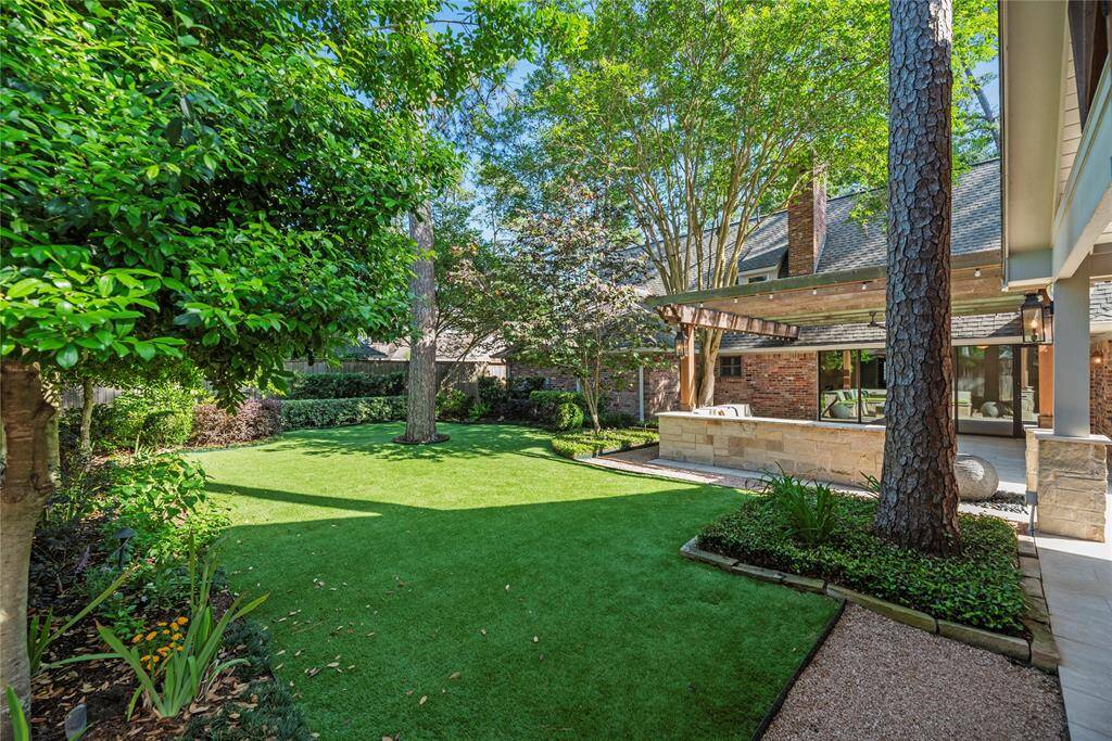 Manicured backyard showcasing a lush synthetic turf, mature trees, and a view of the pergola-covered patio tucked behind the home.