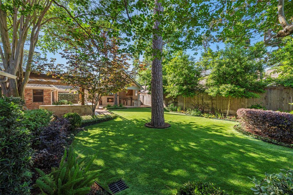 A serene, shaded garden view with carefully manicured beds, blooming foliage, and an open lawn leading to the outdoor fireplace and entertaining area.