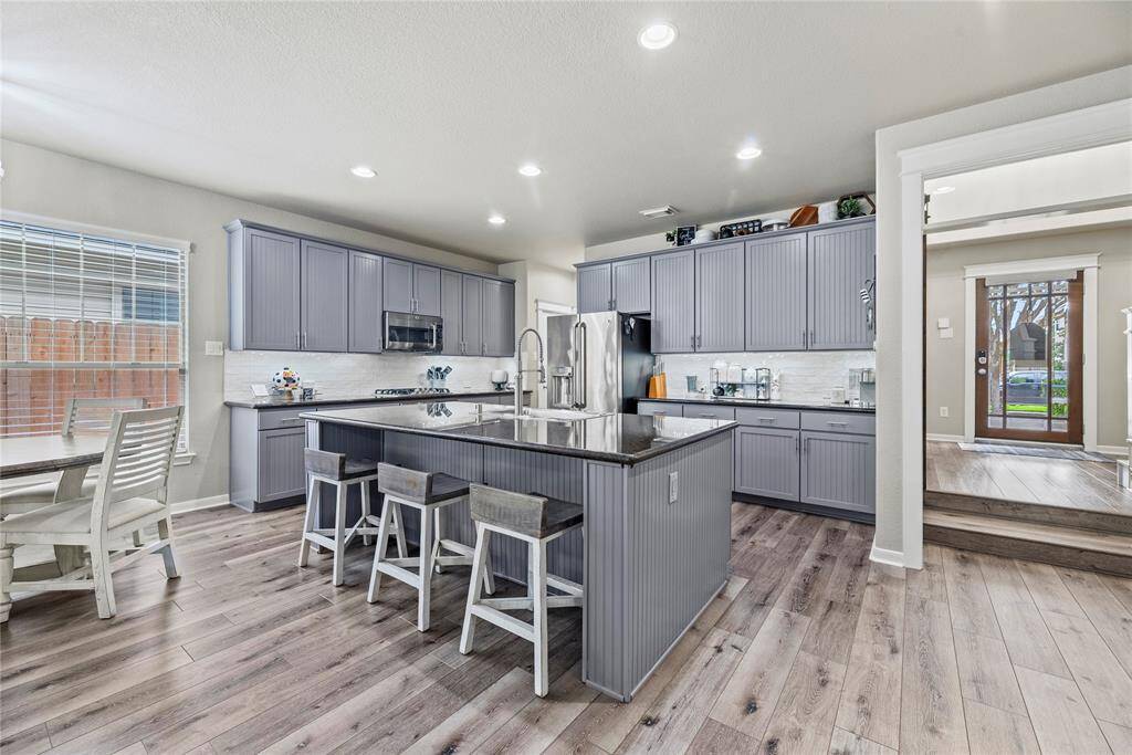 This kitchen features modern gray cabinetry, a large central island with a farmhouse styled sink with seating, stainless steel appliances, granite countertops and under cabinet lighting. The open layout includes the dining area while ample natural light enhances the space.