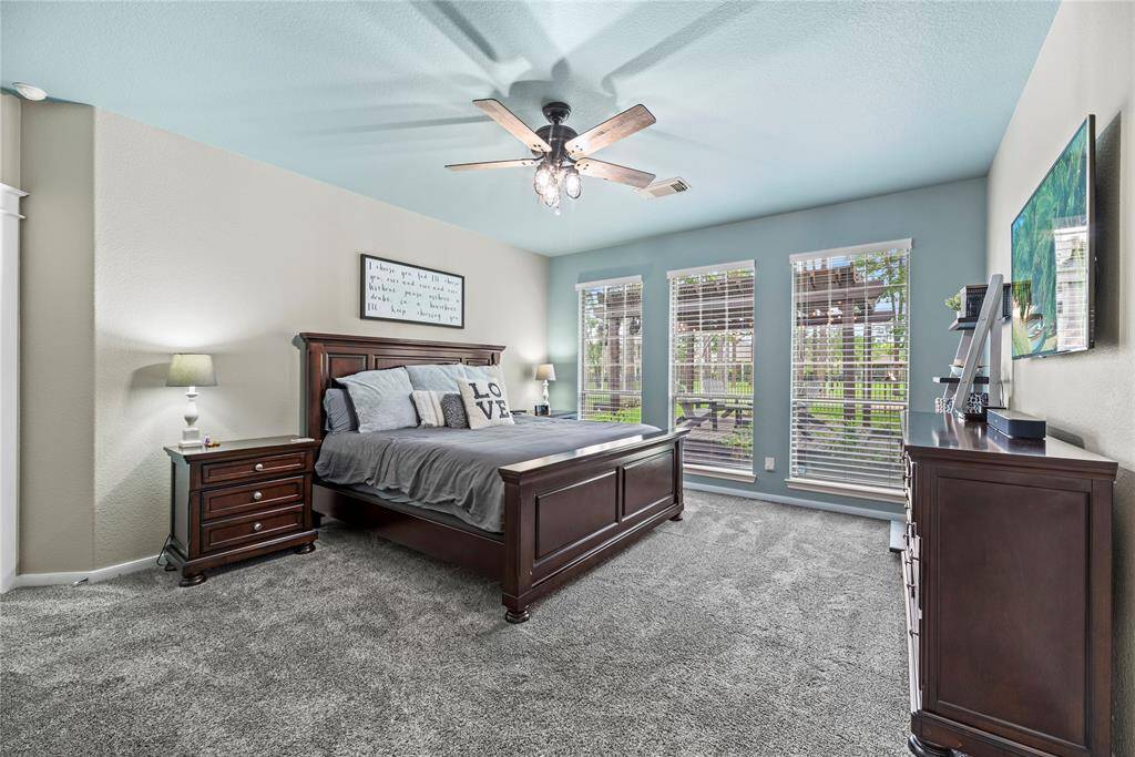 This spacious bedroom features a cozy atmosphere with plush carpeting, a stylish ceiling fan, and abundant natural light from three large windows that overlook the expansive backyard and park and trail behind the home.