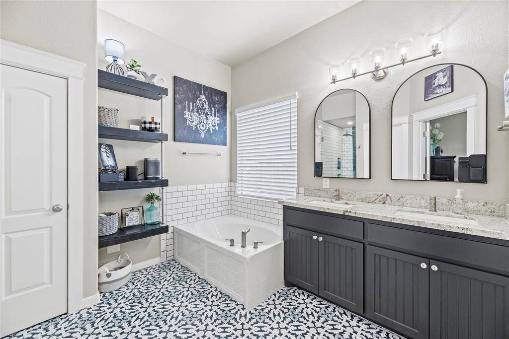 This completely remodeled bathroom features a modern design with custom decorative tile flooring, a dual-sink vanity with granite countertops, and large framed mirrors. It includes a spacious bathtub with subway tile surround, open shelving for storage, and stylish lighting fixtures, creating a bright and elegant space. There is a beautiful shower across from this area that is pictured next.