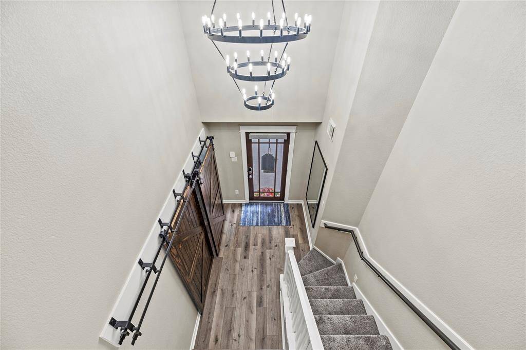 This photo showcases a bright, two-story entryway with a modern chandelier, LPV flooring, and a stylish barn door. The staircase features carpeted steps and a sleek railing, leading to an inviting front door with glass panels. Let's go take a look upstairs!