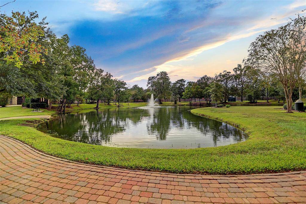 This serene photo showcases a picturesque Coles Crossing pond with a central fountain, surrounded by lush greenery and mature trees. A charming brick pathway borders the water, perfect for leisurely strolls, adding to the tranquil ambiance of this outdoor space.