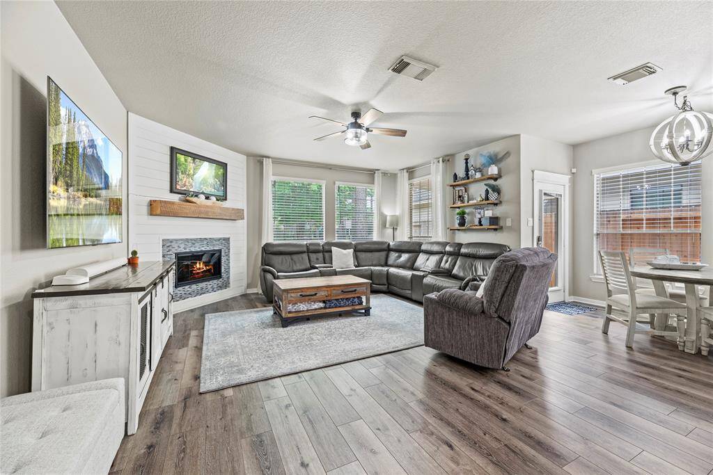 The living room features a gas log fireplace with a wooden mantle, ship lap surround and stylish built-in shelves. The space is brightened by multiple windows, and a modern ceiling fan adds a contemporary touch. Open concept living area with an adjacent dining area with a chic light fixture, all set on elegant LPV flooring.