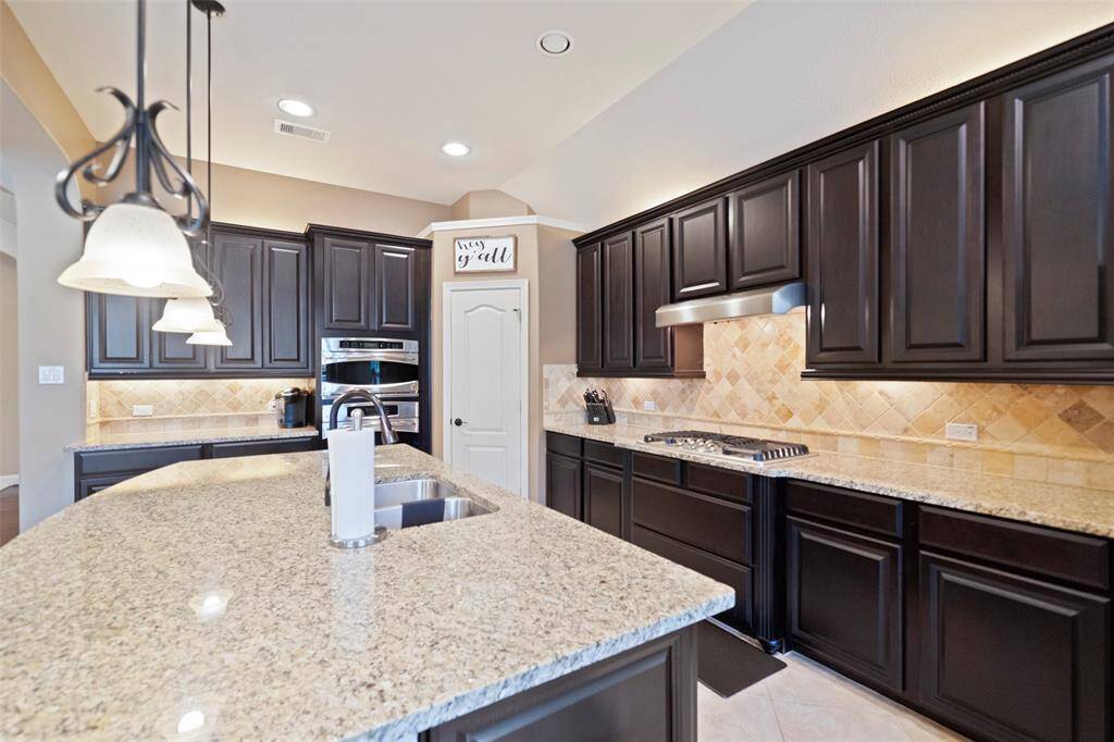 Stunning gourmet kitchen boasts rich espresso cabinetry, sleek granite countertops, stainless steel appliances, and a spacious center island with dual sinks. Under-cabinet lighting and a tumbled stone backsplash add warmth and elegance.