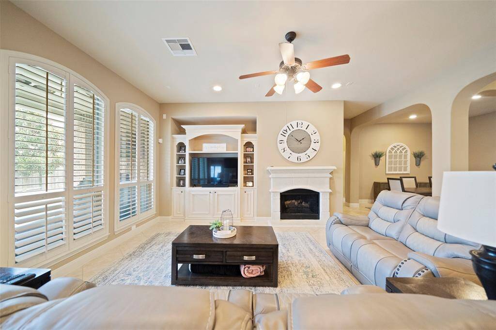Elegant living room with a gas fireplace, custom built-ins, and oversized plantation-shuttered windows offering abundant natural light. A ceiling fan and recessed lighting enhance comfort and style.
