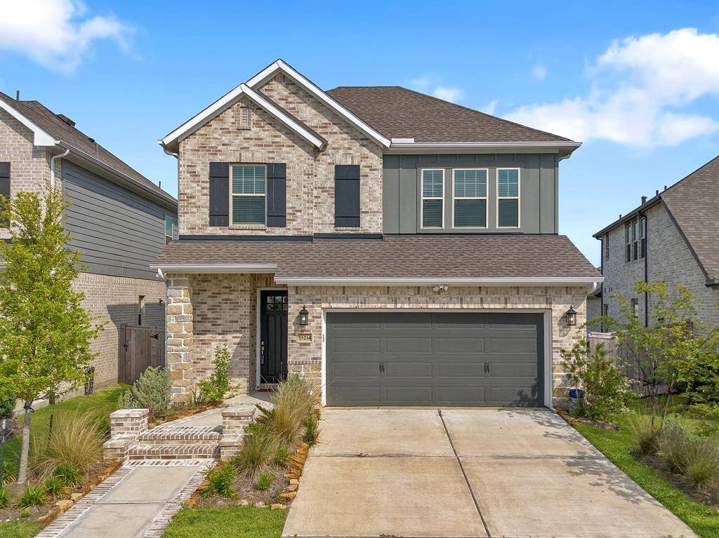 The house features a two - story design with a combination of brick and siding exterior. A spacious driveway leads to a two-car garage adorned by landscaped flower beds.