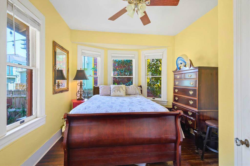 This cheerful secondary bedroom is filled with natural light, thanks to its charming bay windows that offer serene views of the outside garden area. Original hardwood floors and soft yellow walls create a warm, inviting atmosphere—ideal for guests or a cozy reading room and home office