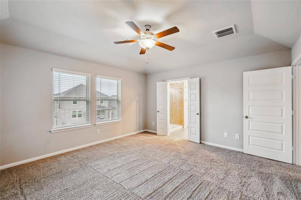 This is a bright, spacious bedroom with neutral walls and carpet, featuring a ceiling fan and two large windows. It includes an en-suite bathroom accessible through double doors.