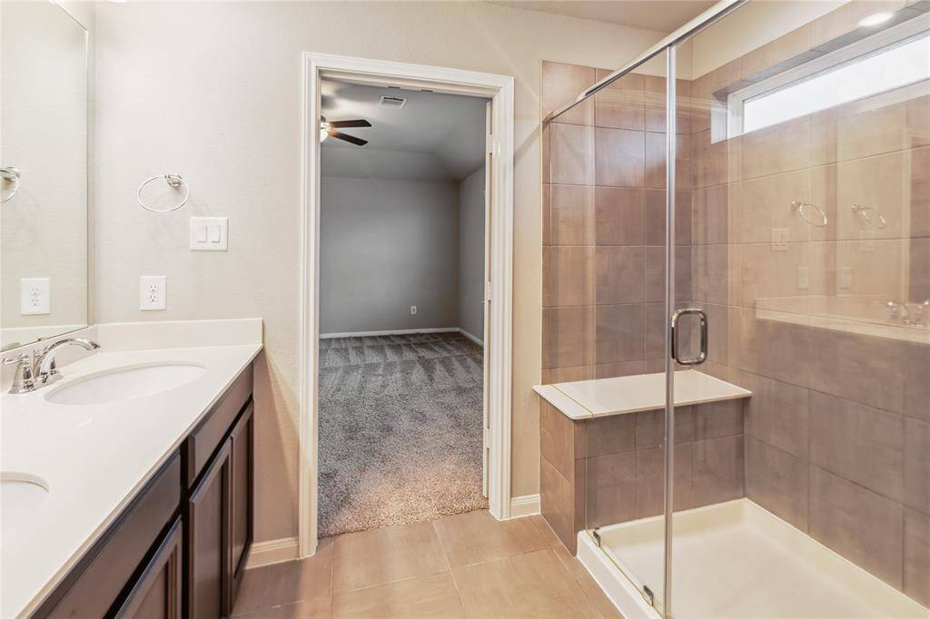 Modern bathroom with a double sink vanity, sleek countertops, and a large glass-enclosed shower. The room is well-lit and leads into a carpeted bedroom area.