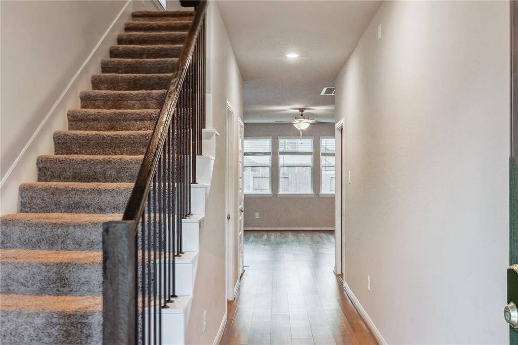 Welcoming entryway with a carpeted staircase on the left and a long hallway leading to a well-lit room with hardwood floors and large windows.