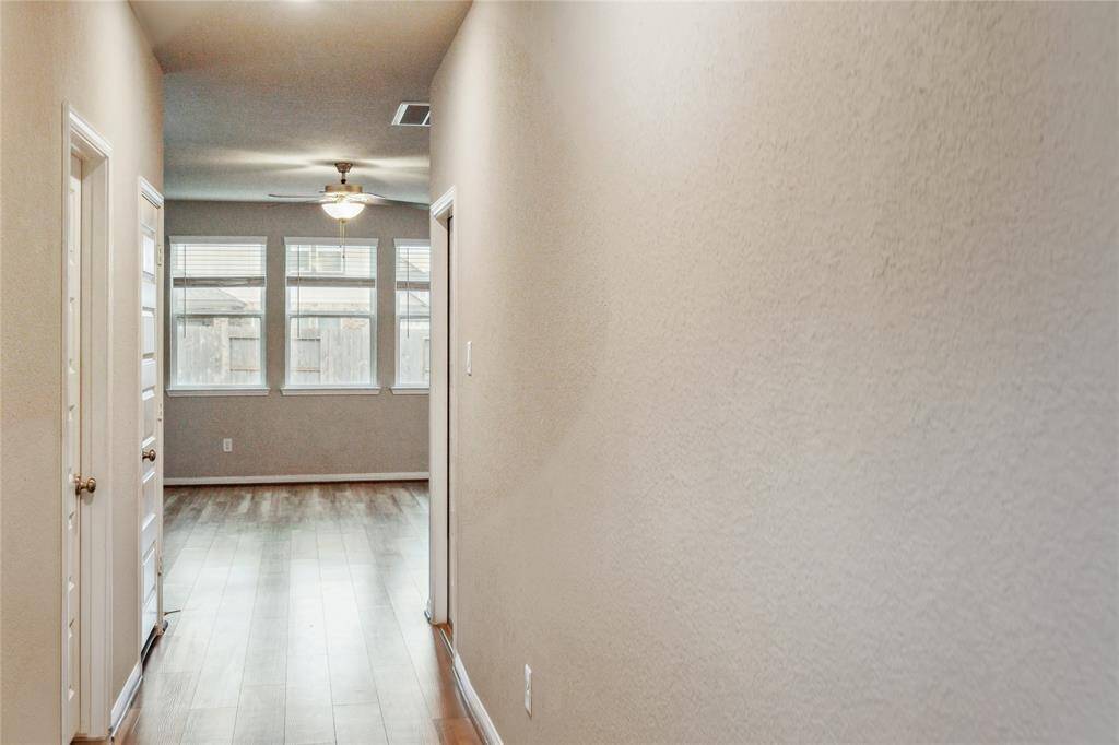 Hallway with light walls leading to a bright room with large windows and wood flooring. The room features a ceiling fan and provides ample natural light.