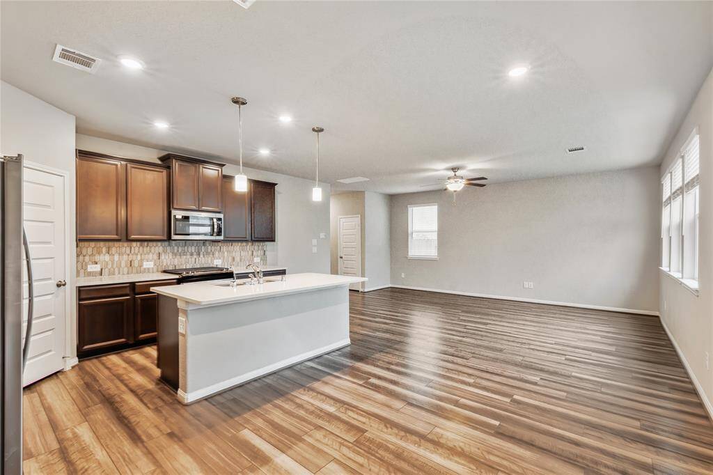 Spacious open-concept kitchen and living area. The kitchen features dark wood cabinets, a modern backsplash, and a large island with pendant lighting. The living area has polished wood flooring, ample natural light from multiple windows, and a ceiling fan for comfort.