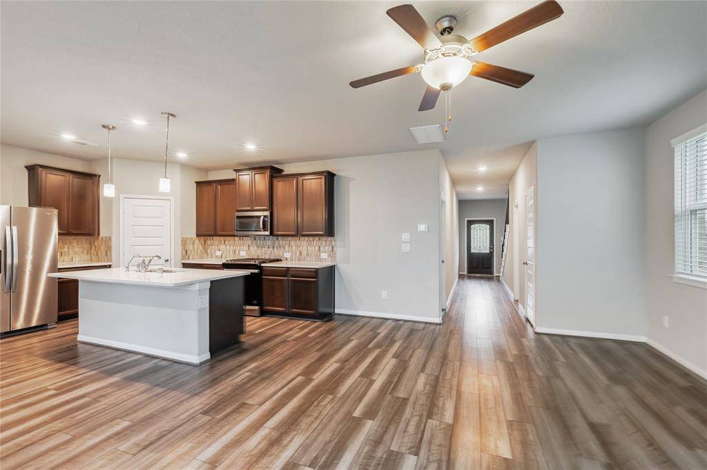 Showcases a modern, open-concept kitchen and living area with hardwood floors. The kitchen features dark wood cabinets, stainless steel appliances, a central island, and pendant lighting. There's a ceiling fan in the living area and a hallway leading to other rooms. Abundant natural light fills the space through multiple windows.