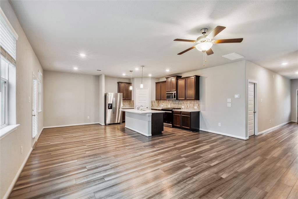 This open-concept living area features modern wood-like flooring and a sleek kitchen with dark cabinetry, stainless steel appliances, and a central island. The space is brightened by recessed lighting and a ceiling fan, creating a welcoming atmosphere.