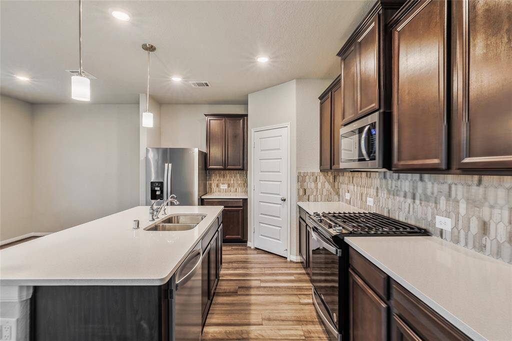 This modern kitchen features dark wood cabinetry, a sleek island with a built-in sink, and stainless steel appliances, including a fridge and oven. The backsplash adds a stylish touch, complemented by warm wood flooring and recessed lighting.