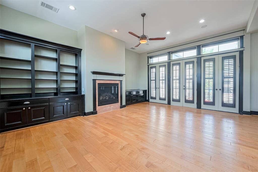 Airy and welcoming, the living room features a fireplace framed by custom shelving, illuminated by recessed lights and a ceiling fan. Three sets of glass-paneled doors topped with transom windows invite light in and open to the balcony beyond.