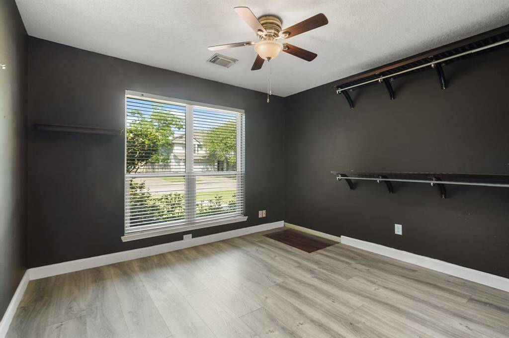 This bedroom features dark walls, light wood vinyl floors, and a large window with blinds, allowing natural light to fill the space. It includes wall-mounted shelves and a ceiling fan with a light fixture, offering functionality and comfort.