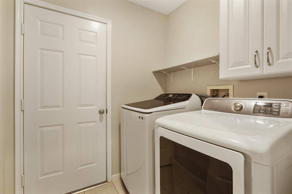 This photo shows a laundry room with white cabinetry, featuring a washer and dryer next to a storage cabinet and a small shelf above. Door leads to 2 car garage.