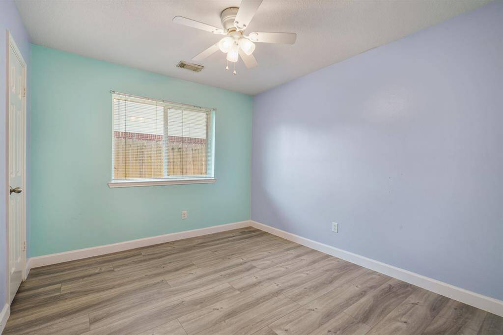 This bedroom features light blue walls, a ceiling fan, and wood-style vinyl flooring. It has a window with blinds for natural light and a simple, clean design.