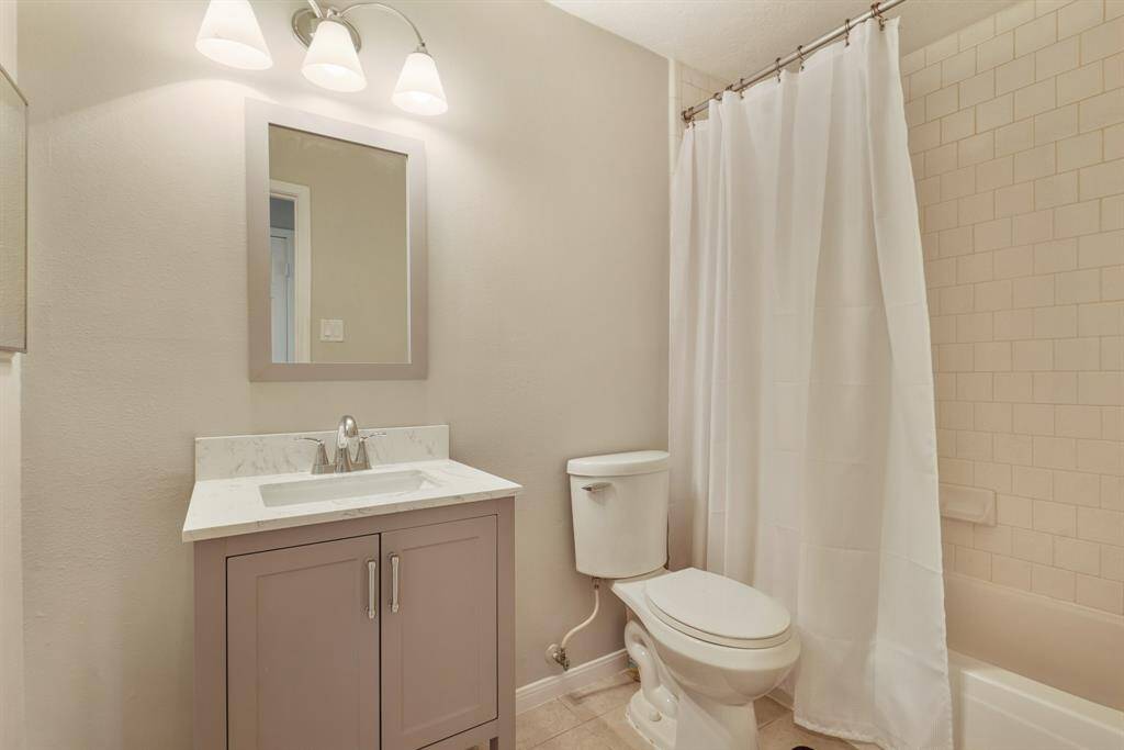 This is a modern bathroom featuring a sleek vanity with a large mirror, and three-light fixture. It includes a toilet and a bathtub shower combo with a tiled wall.