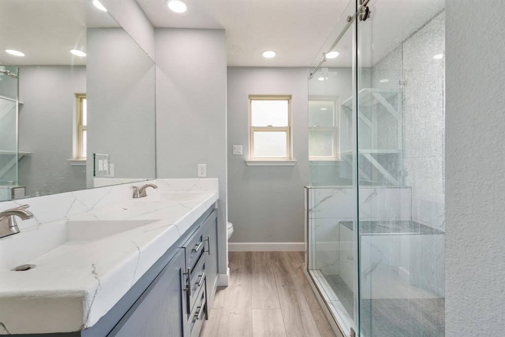 This modern bathroom features a sleek double vanity, a spacious glass-enclosed shower, and elegant wood vinyl flooring. Soft, neutral tones create a calming atmosphere, complemented by recessed lighting and a small window for natural light by the toilet.