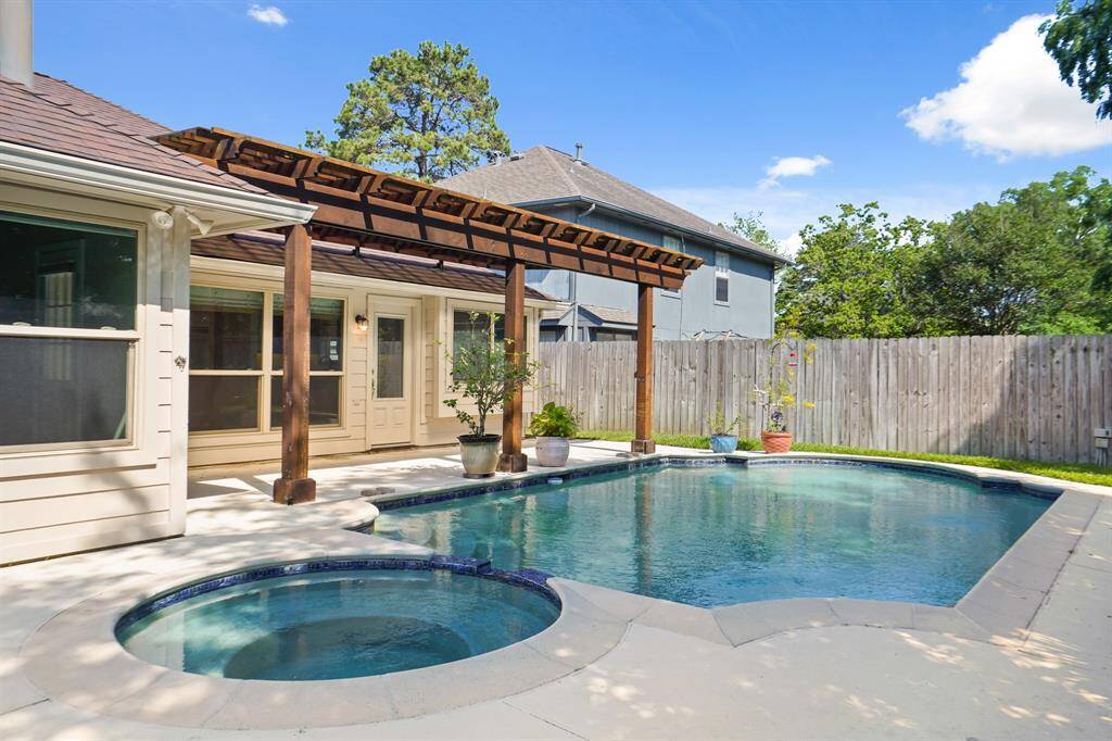 This backyard features a beautiful swimming pool with an attached hot tub, surrounded by a spacious patio. A wooden pergola provides partial shade, and the area is fenced for privacy, creating a serene outdoor retreat.