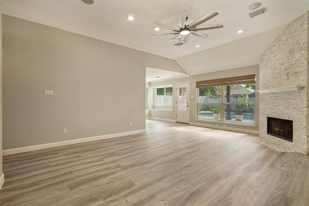 This spacious living room features light wood vinyl flooring, a modern ceiling fan, and a stone fireplace. Large windows and a back door provide ample natural light, leading to a view of the backyard.