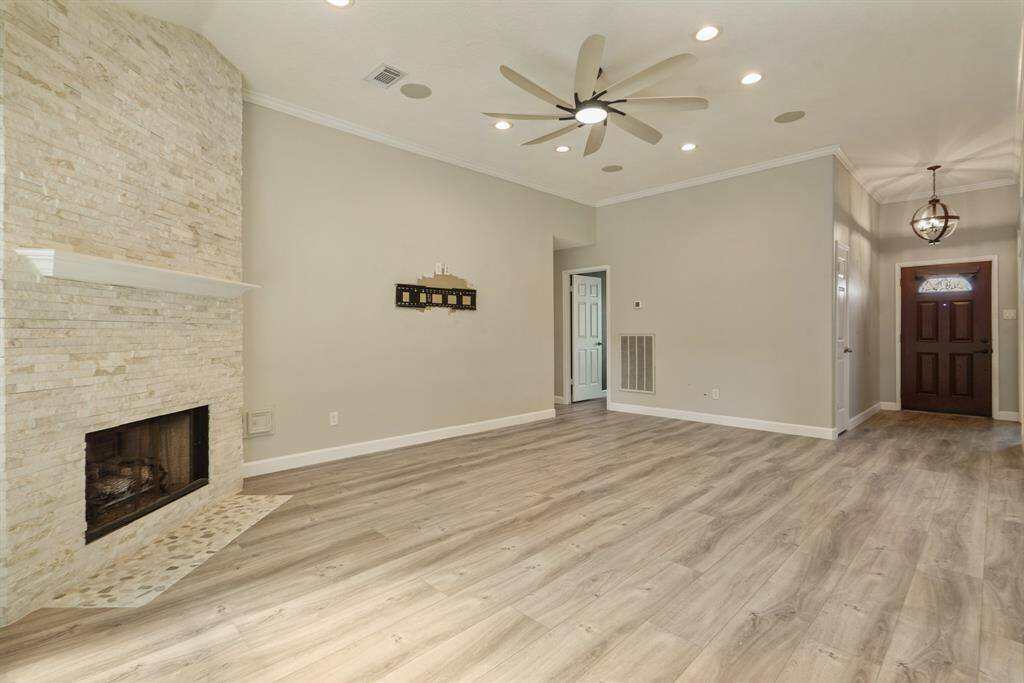 This spacious, living room features a modern ceiling fan, recessed lighting, and a cozy stone fireplace. The light wood flooring complements the neutral walls, offering a warm and inviting atmosphere. A stylish light fixture hangs near the entrance, adding charm to the entryway.
