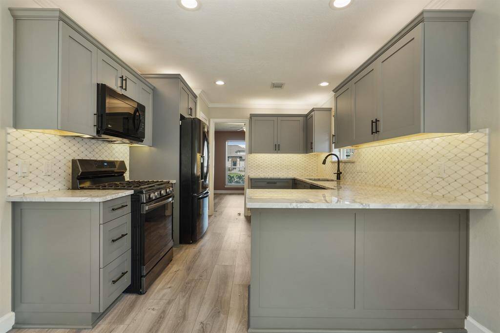 This updated kitchen features modern cabinetry in a soft gray tone, with sleek black appliances, a stylish tile backsplash, and under-cabinet lighting. The space is well-lit and boasts a functional layout with ample countertop space.