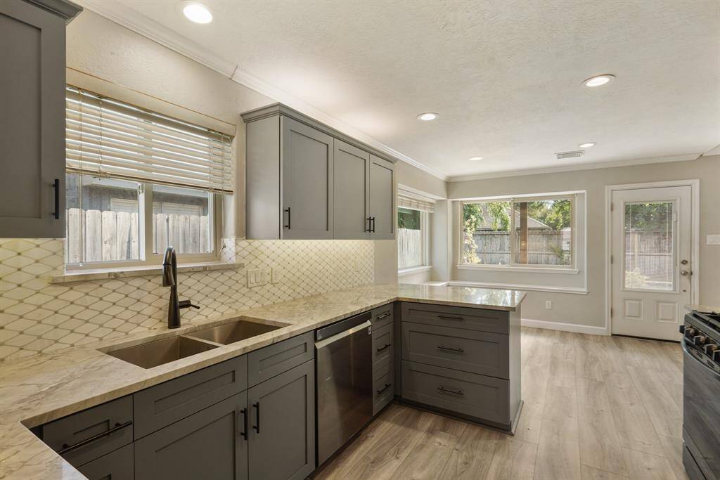 This updated kitchen is modern and spacious, featuring sleek gray cabinets, marble countertops, and a stylish tile backsplash. It includes stainless steel appliances, recessed lighting, and large windows providing natural light and views of the backyard.
