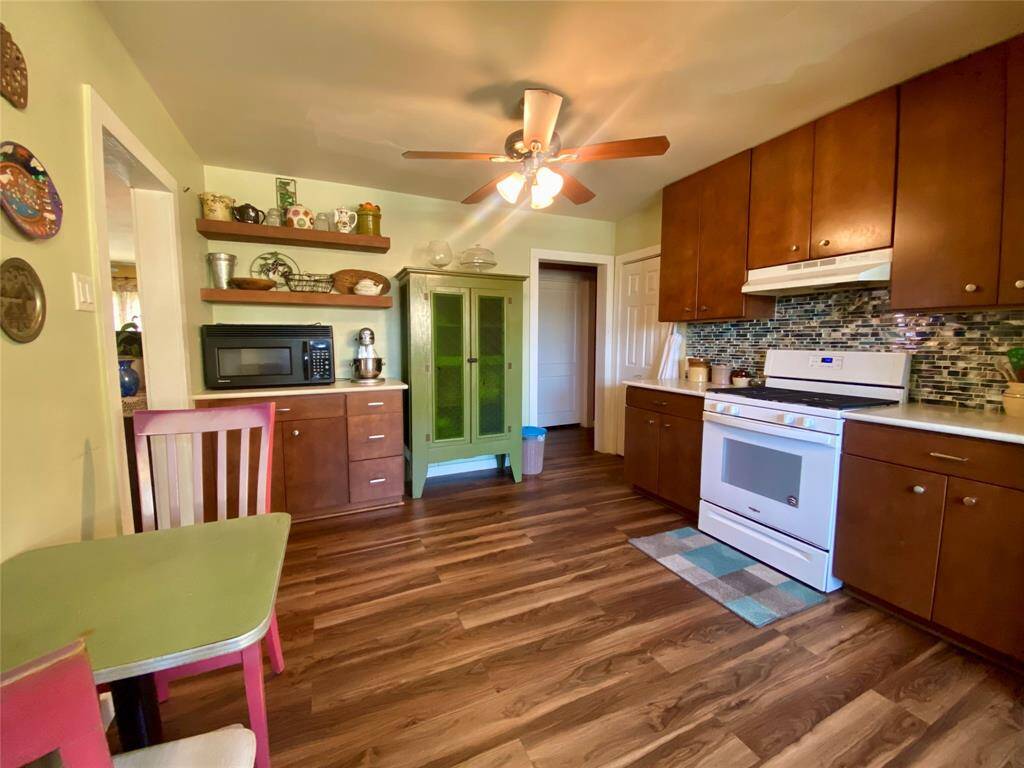 Charming kitchen with warm wood cabinets, a modern backsplash, and wood flooring. Features a ceiling fan, white appliances, and open shelving for decor. Cozy dining area with colorful accents.