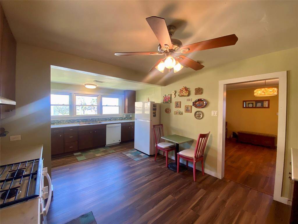 Another view of the kitchen with warm lighting, featuring a ceiling fan, wood flooring, and a quaint dining nook. The space is adorned with wall decor and includes a stove, refrigerator, and ample countertop space with large windows for natural light.