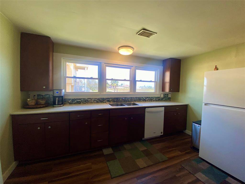 This kitchen features ample natural light from large windows, wood cabinets, and a modern backsplash. It includes essential appliances like a refrigerator and dishwasher, with wood flooring for a warm touch.