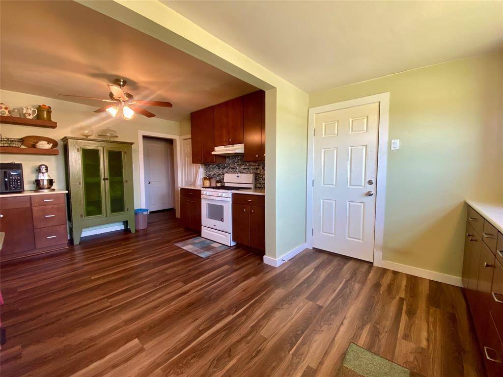 This kitchen features warm wood flooring, a classic white stove, and ample cabinetry for storage. A ceiling fan provides ventilation, and the room is bright and inviting with a light green accent wall. The door to the right leads to the utility room with back door and deck access.