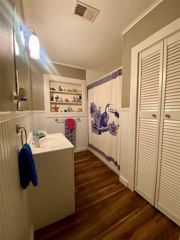 Guest bathroom features a cozy design with wood-style flooring and soft lighting. It has a vanity with a mirror, built-in shelving for storage, and linen closet with louvered doors. The walls are painted in a neutral color, complemented by wainscoting for added charm.