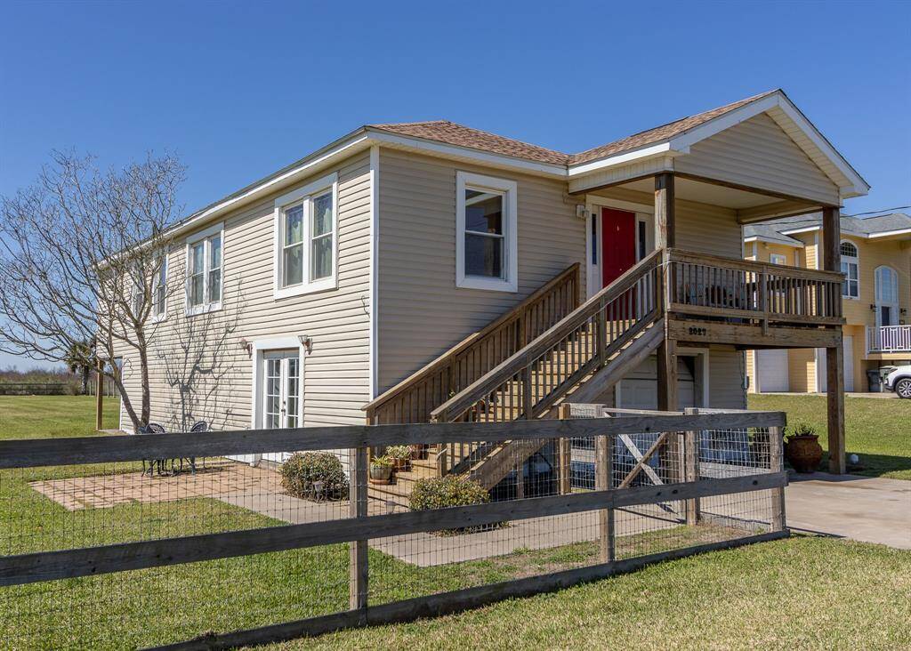 This charming two-story home features a beige exterior with a covered porch and red door. A wooden staircase provides access to the main entrance. It has a spacious, fenced yard and a patio area, perfect for outdoor activities.